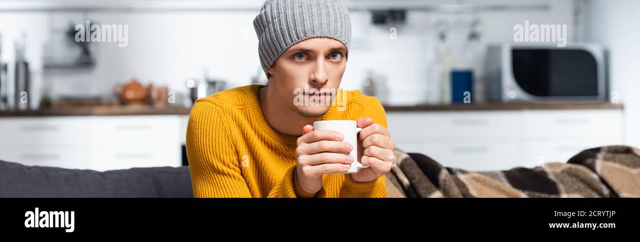 orientamento panoramico del giovane in maglia maglione e cappello guardando la fotocamera mentre si tiene una tazza di bevanda calda Foto Stock