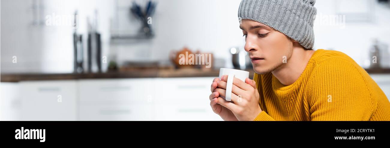 intestazione del sito web di giovane uomo in maglia cappello tenendo tazza di bevande calde in cucina fredda Foto Stock