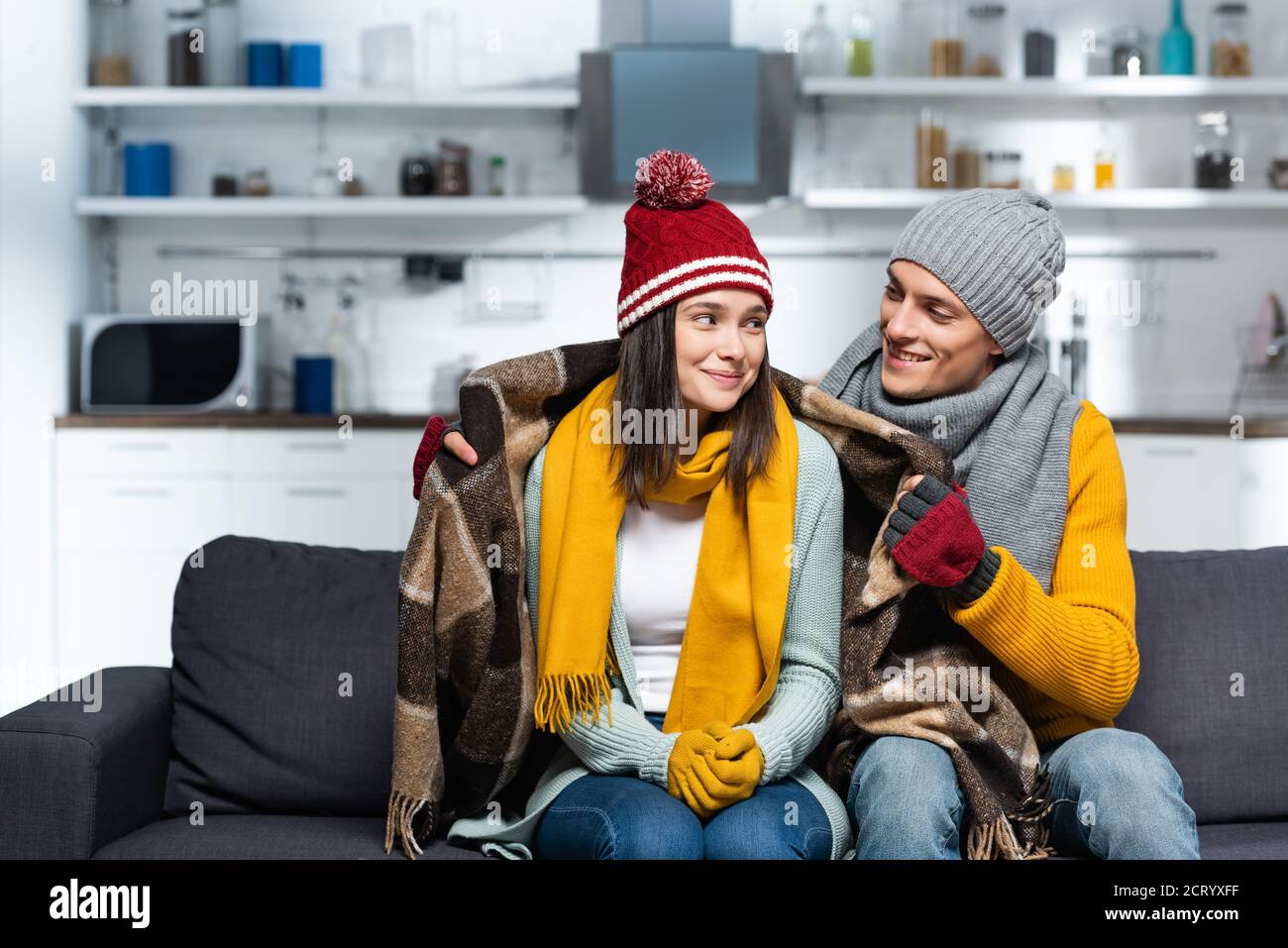 giovane uomo in cappello caldo e guanti che coprono la ragazza gelida con  una coperta di plaid mentre si siede sul divano in cucina fredda Foto stock  - Alamy
