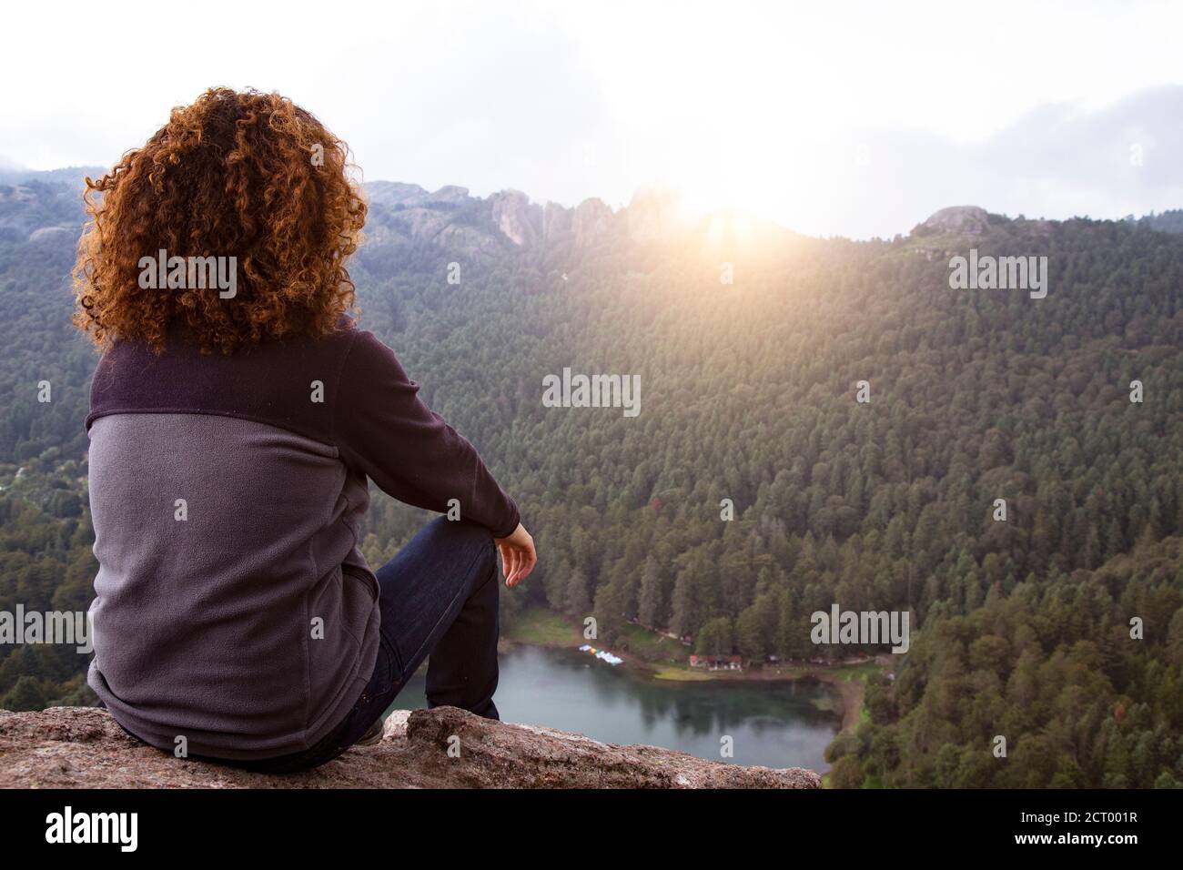 Escursionista maschile con capelli ricci, seduto sul bordo della scogliera, ammirando l'alba Foto Stock