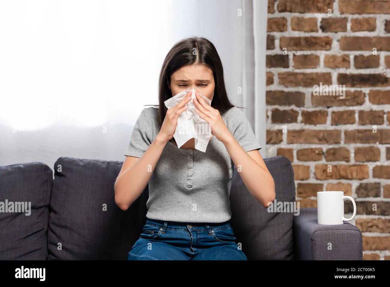 donna malata starnutisce in tovagliolo di carta mentre si siede sul divano vicino a una tazza di bevanda calda Foto Stock