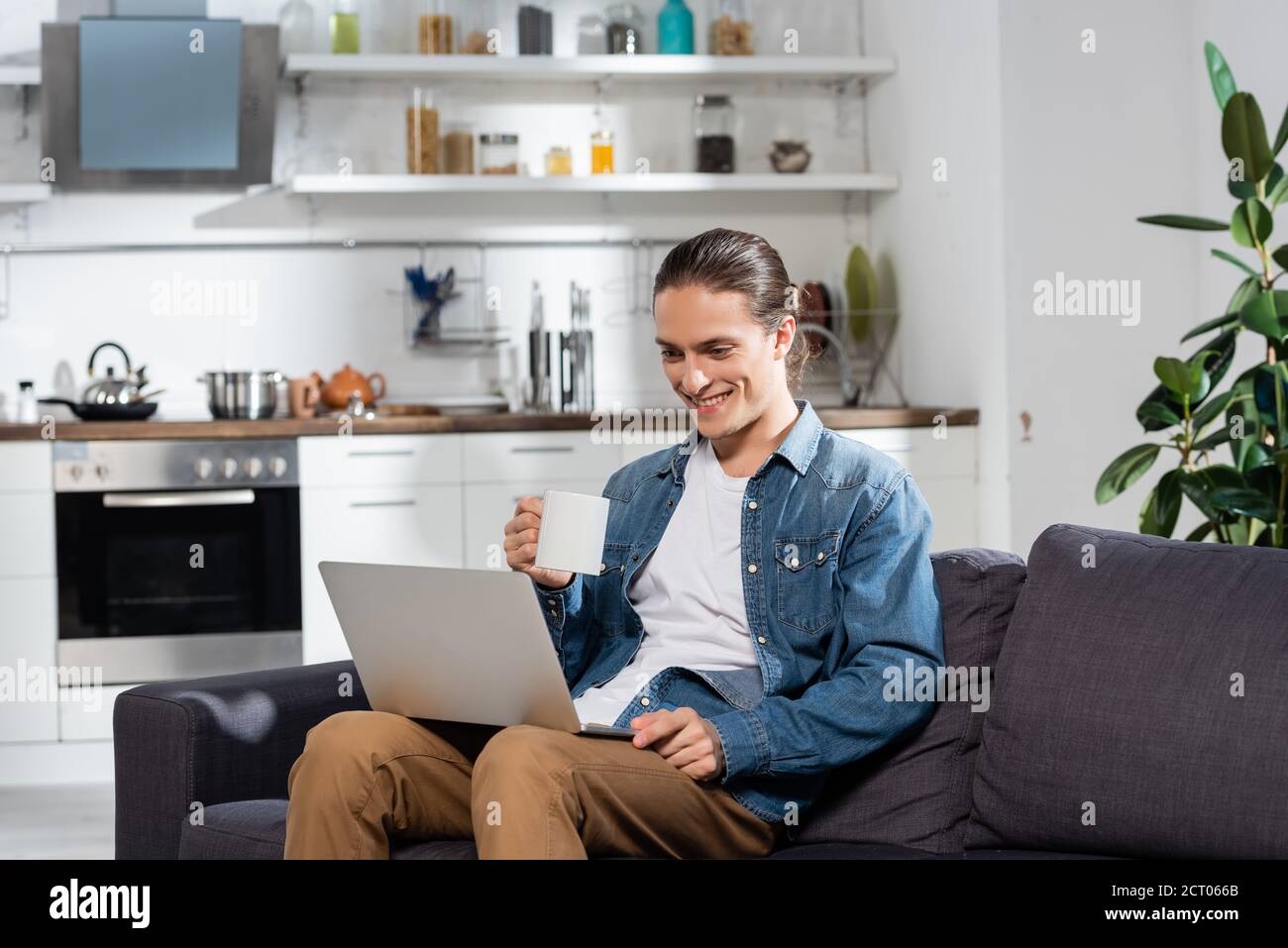 giovane freelance che tiene una tazza di tè mentre si siede sul divano in cucina e utilizzando il computer portatile Foto Stock
