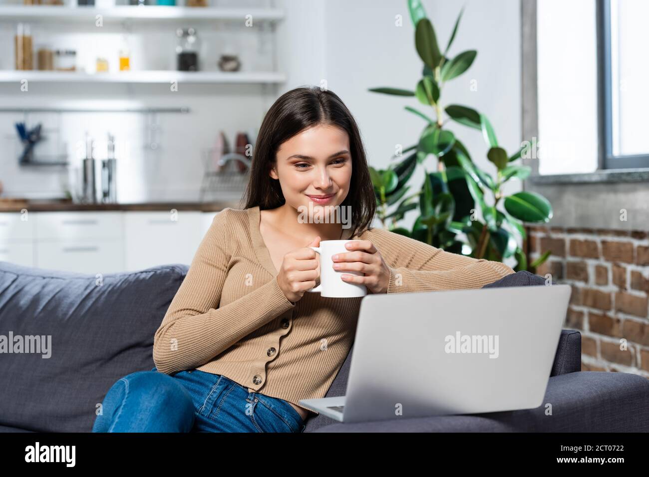 un'allegra freelance che guarda il computer portatile mentre si siede sul divano cucina con tazza di bevanda calda Foto Stock