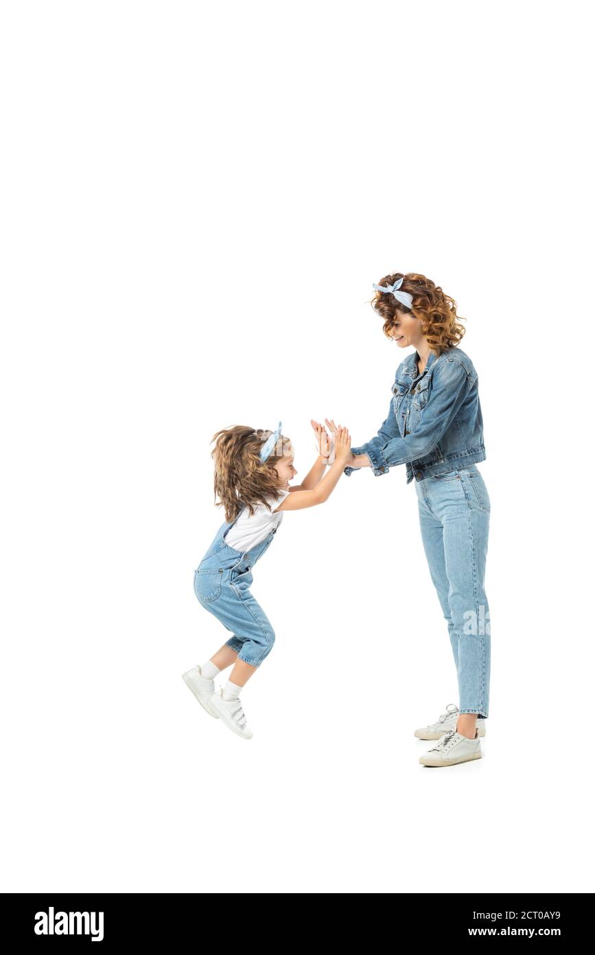 vista laterale della figlia in jeans vestito saltando e aggrappando mani con madre isolata su bianco Foto Stock
