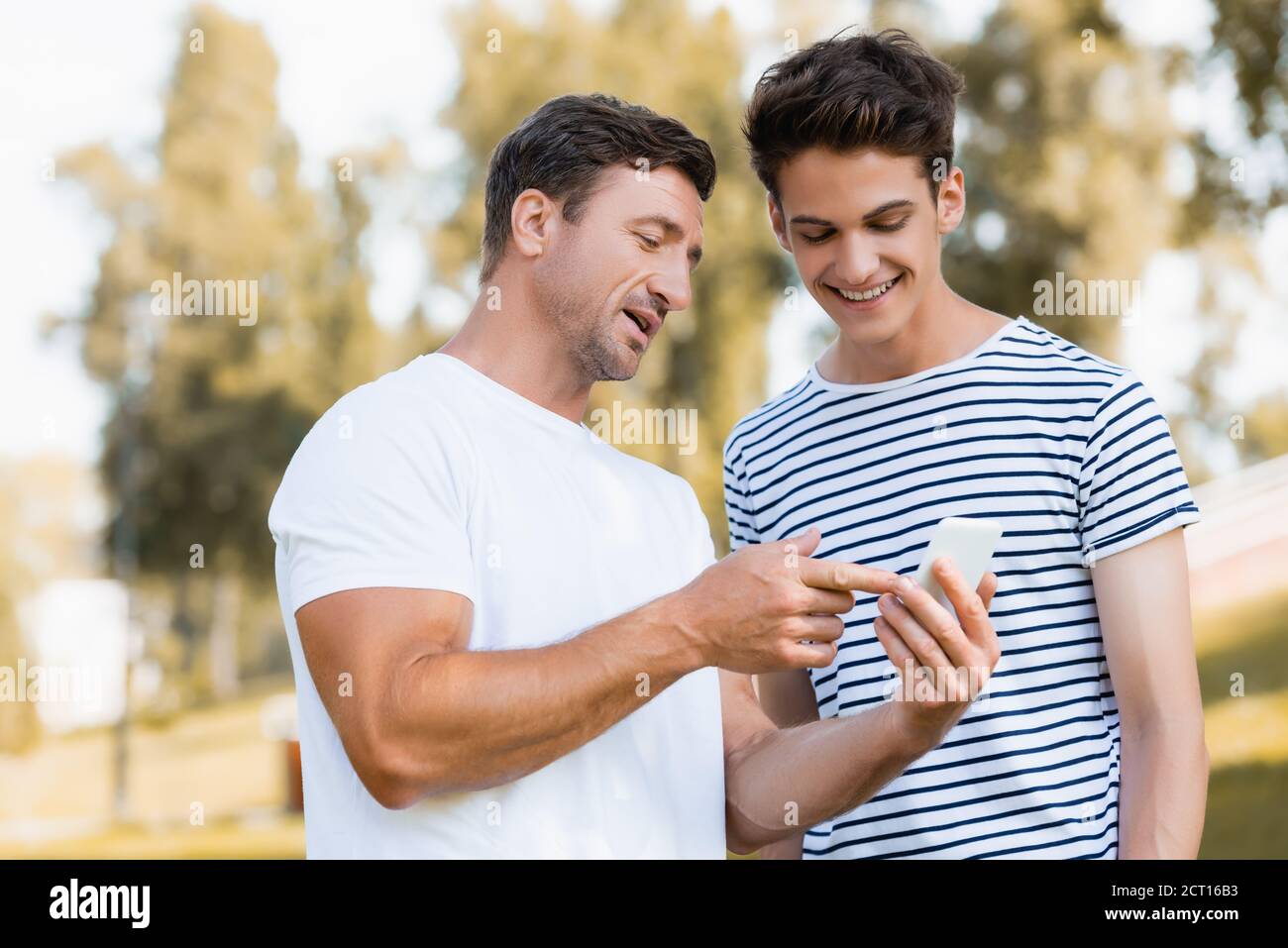 uomo che punta con il dito contro lo smartphone vicino al figlio adolescente parcheggio Foto Stock