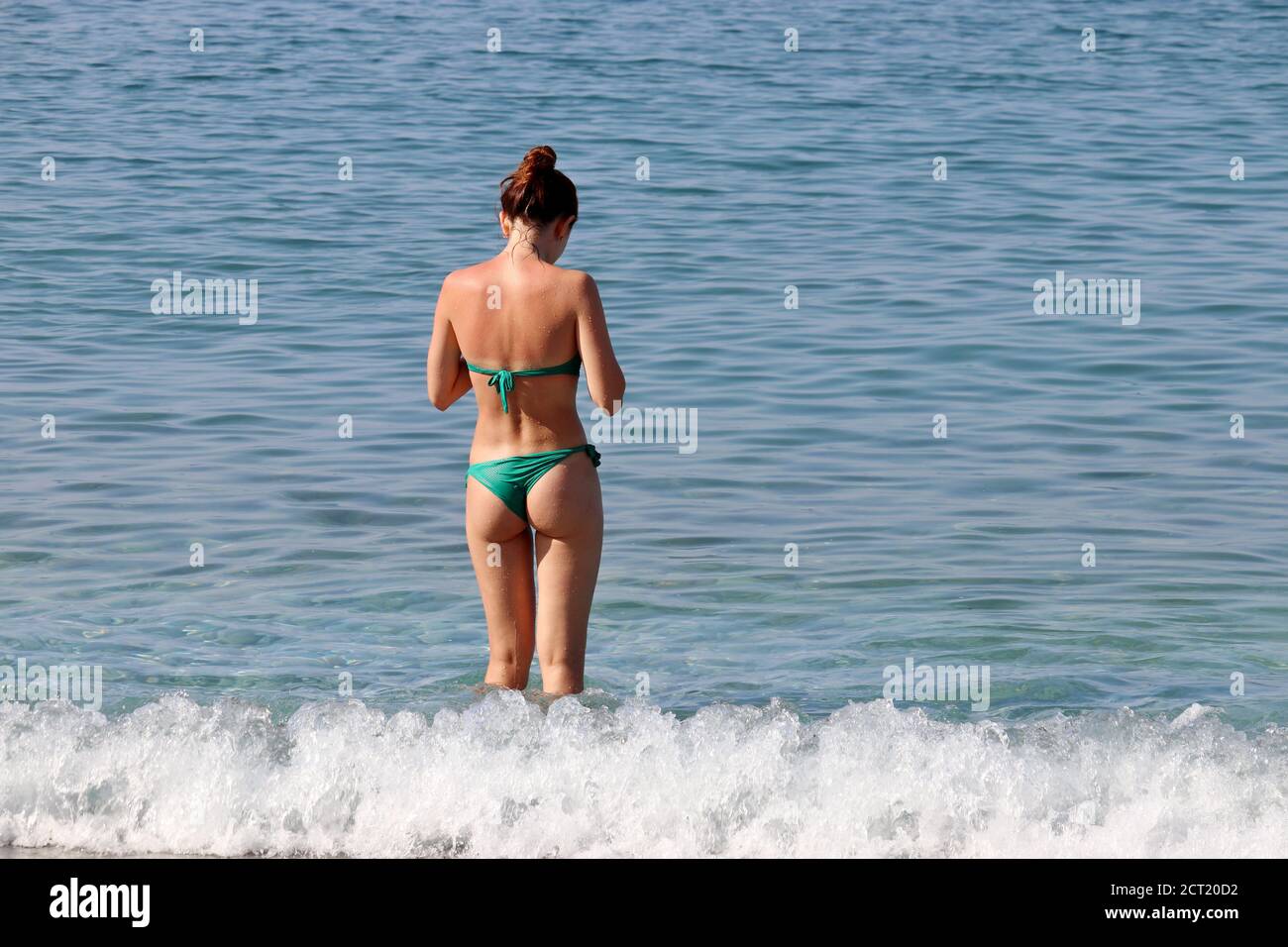 Donna sottile in bikini verde in piedi in onde da surf, vista posteriore. Vacanza in spiaggia, relax e tempo libero sul mare Foto Stock