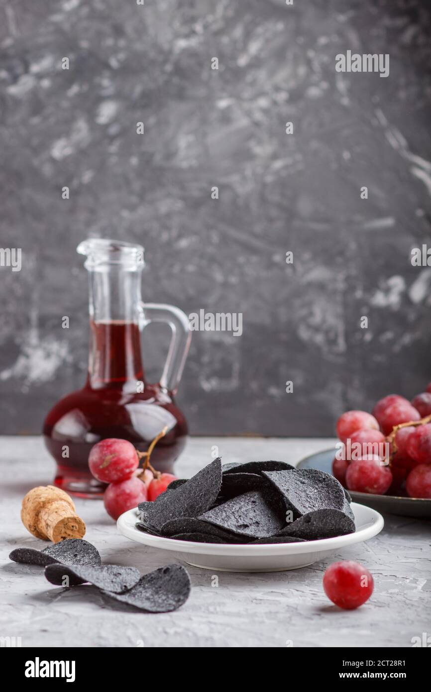 Patatine nere con carbone, aceto balsamico in vetro, uva rossa su un piatto di ceramica blu su fondo grigio. Vista laterale, primo piano, se Foto Stock