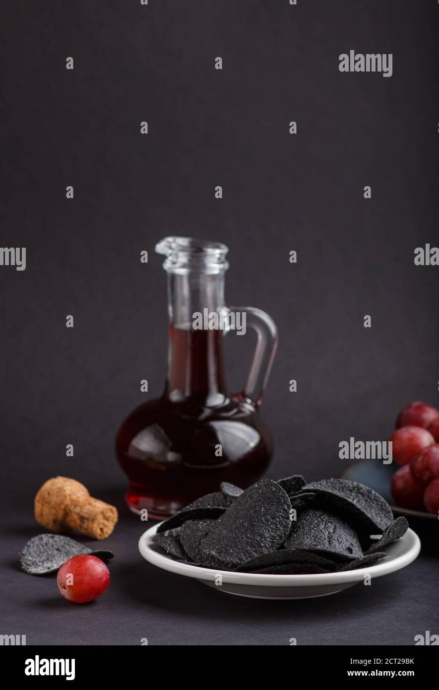 Patatine nere con carbone, aceto balsamico in vetro, uva rossa su un piatto di ceramica blu su fondo nero. Vista laterale, primo piano, selettivo Foto Stock