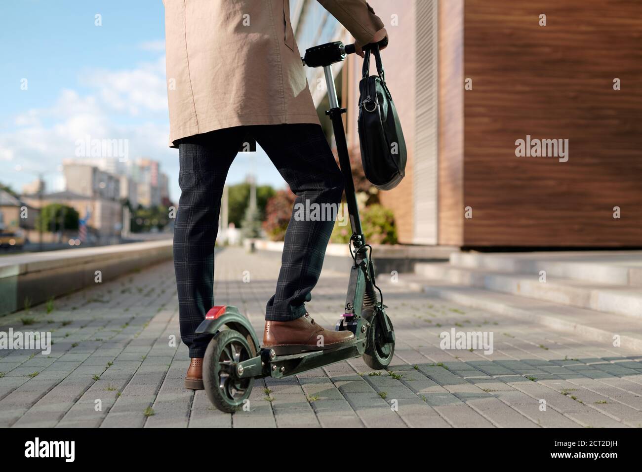 Sezione bassa di uomo d'affari in trenchcoat beige e pantaloni in piedi su scooter Foto Stock