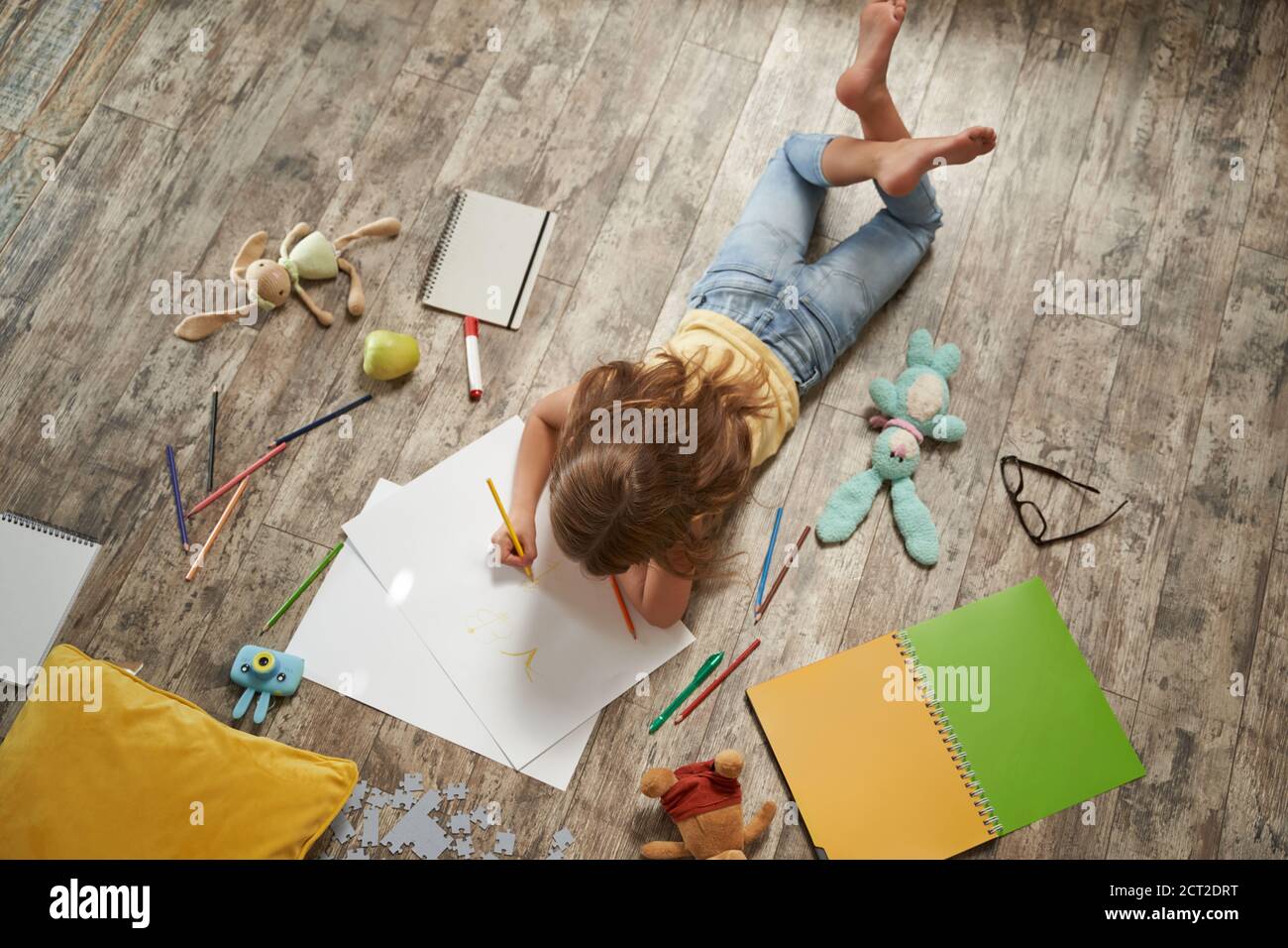Vista dall'alto della bambina caucasica sdraiata sul legno pavimento a casa e disegno con matite colorate su un foglio bianco di carta Foto Stock