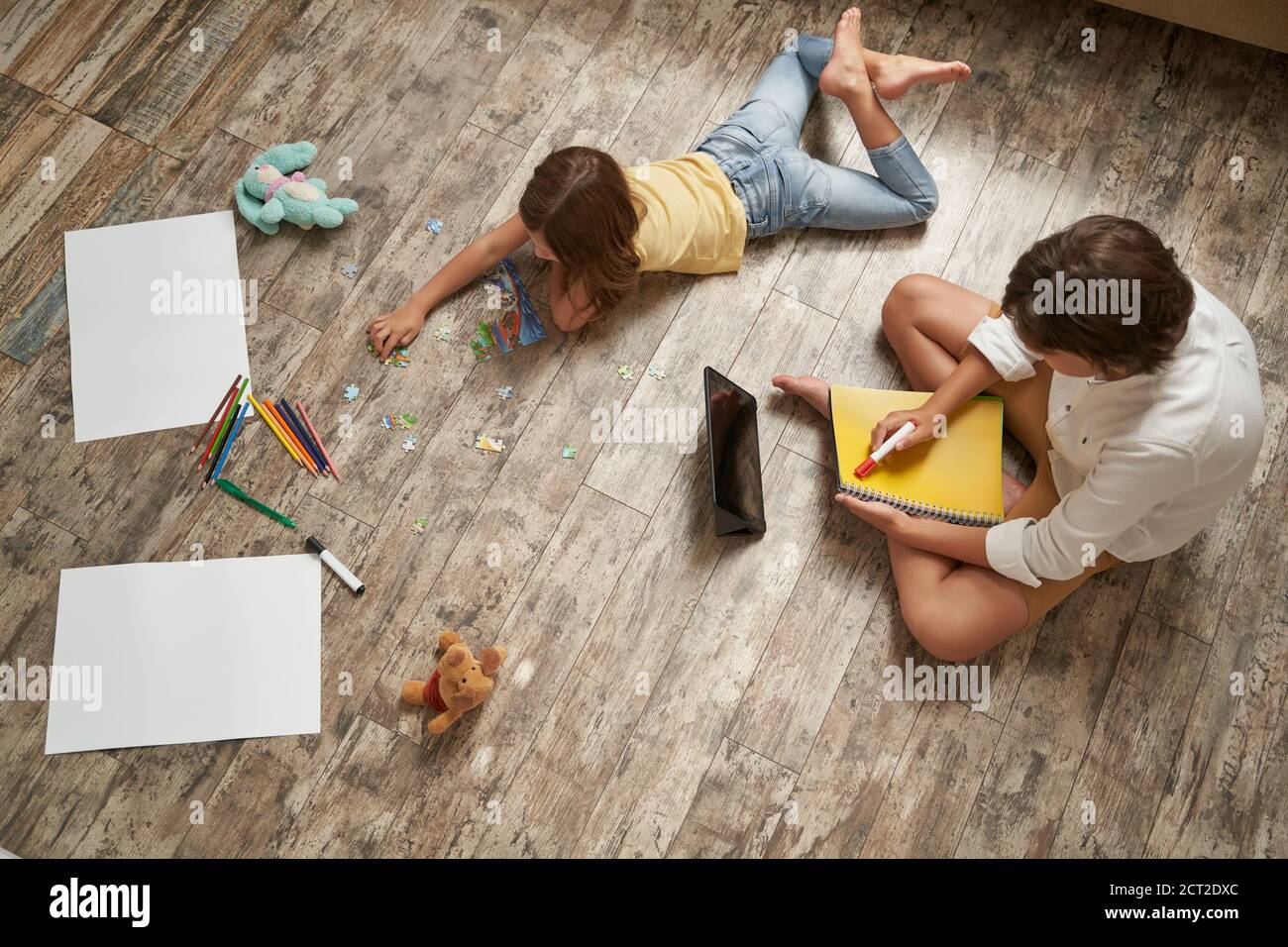 Fratello e sorella giacenti sul pavimento di legno a casa e passare il tempo insieme. Bambina che gioca con puzzle, ragazzo utilizzando tablet digitale e. Foto Stock