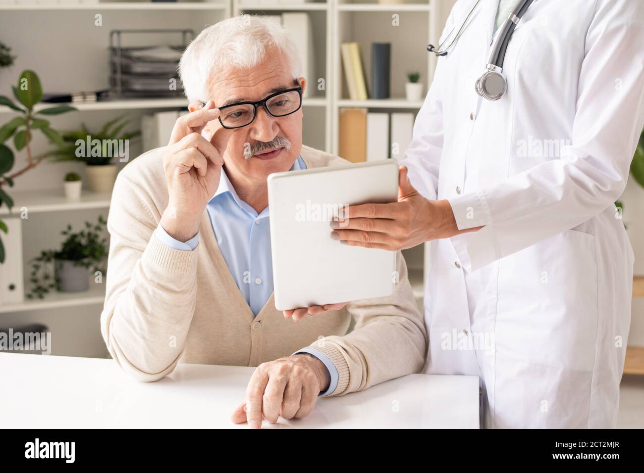 Paziente anziano in occhiali e casualwear guardando i dati su display tablet Foto Stock