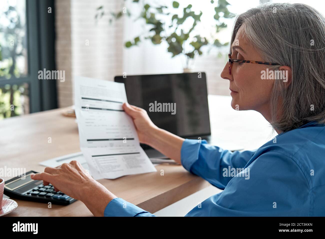 Donna matura che tiene la fattura della carta usando la calcolatrice che gestisce le finanze a casa. Foto Stock
