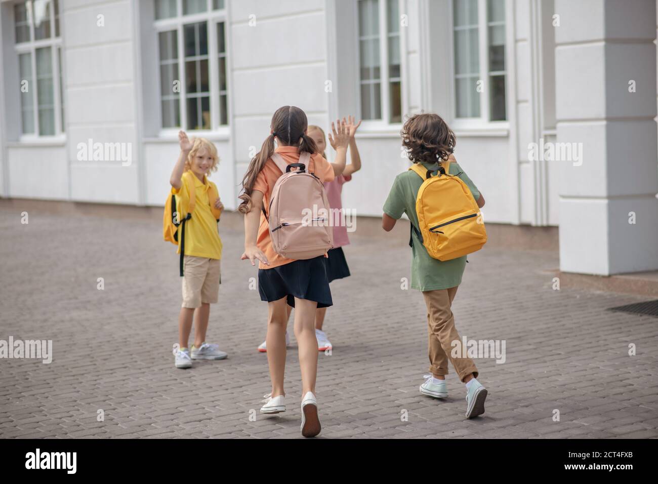 Gli scolari in maschere protettive nel cortile della scuola salutano ciascuno altre mani agitando Foto Stock