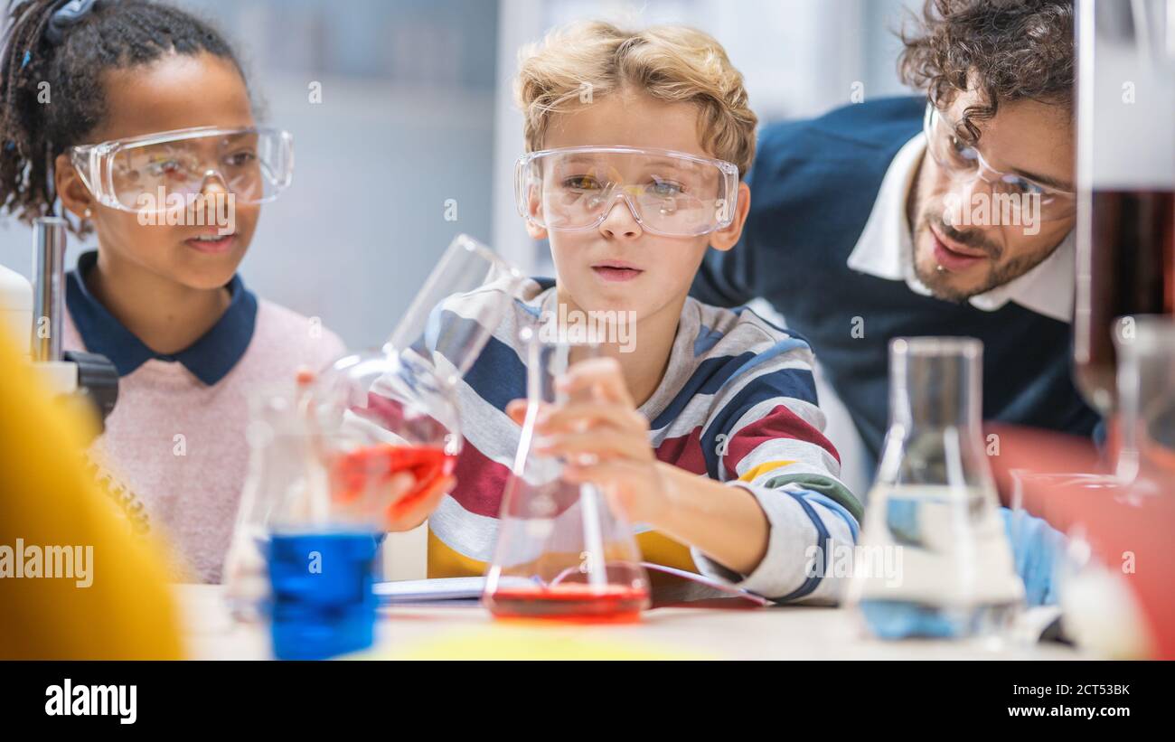 Scuola elementare Scienza Aula: Insegnante entusiasta spiega la chimica a diversi gruppi di bambini, Little Boy Mixes Chemicals in Beakers Foto Stock