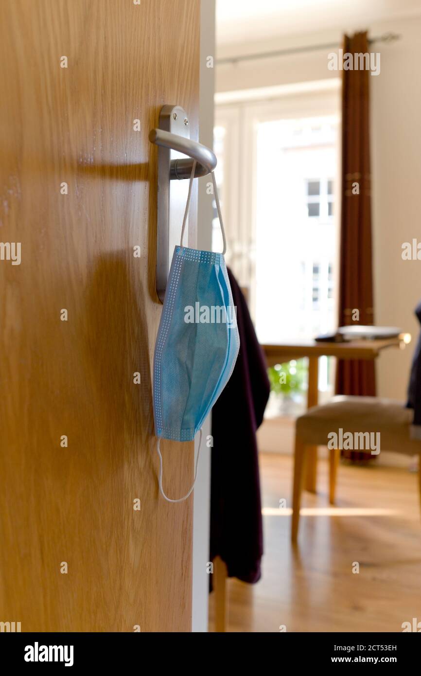 Usato maschera chirurgica usa e getta, appende su una maniglia di porta di metallo su una porta aperta alla sala da pranzo con tavolo in un moderno pulito appartamento dall'aspetto soleggiato Foto Stock