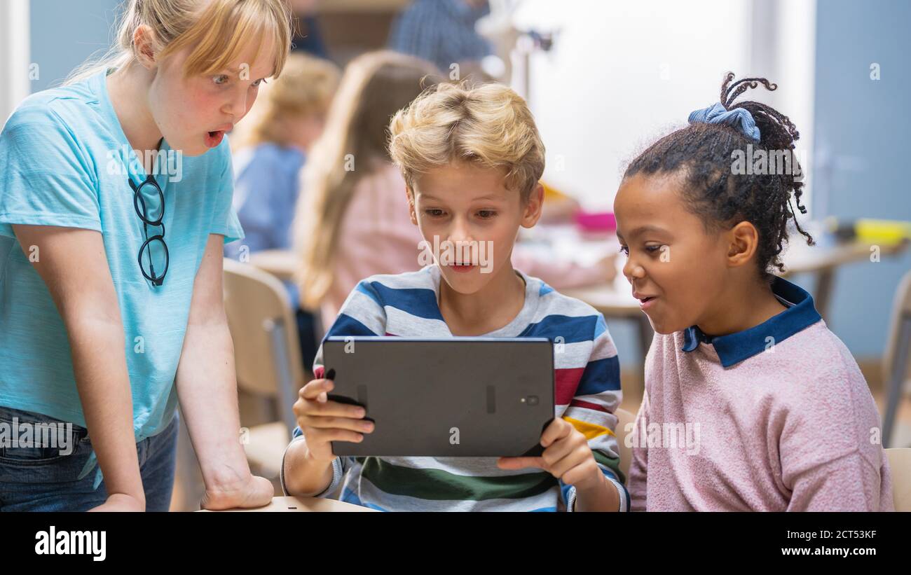 Scuola elementare computer Science Class: Due ragazze e ragazzi utilizzano il Tablet computer digitale con software di realtà aumentata, sono entusiasti, pieno di Foto Stock