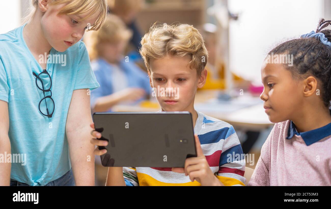 Scuola elementare computer Science Class: Due ragazze e ragazzi utilizzano il Tablet computer digitale con software di realtà aumentata, sono entusiasti, pieno di Foto Stock