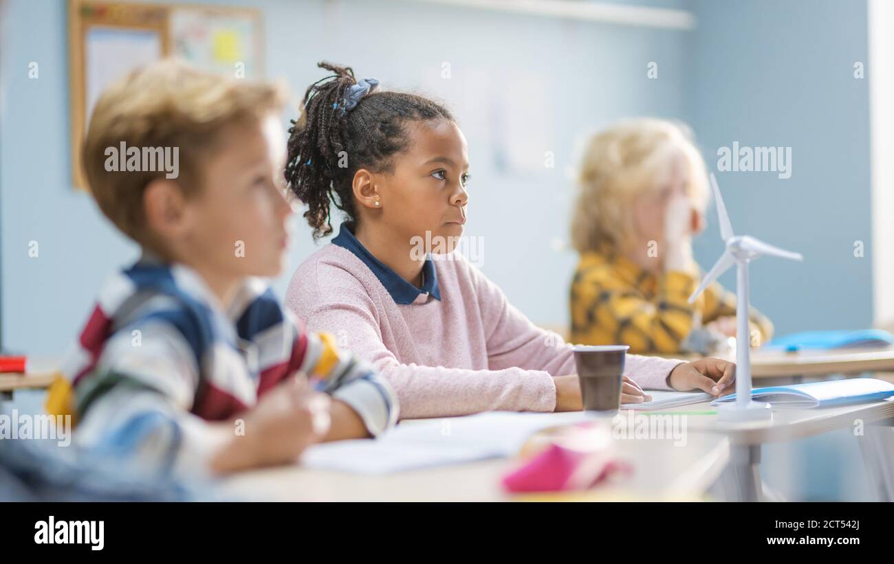 Nella classe della scuola elementare Brilliant Black Girl sta ascoltando con attenzione un insegnante. Junior Classroom con Gruppo di Bright Children che lavorano Foto Stock