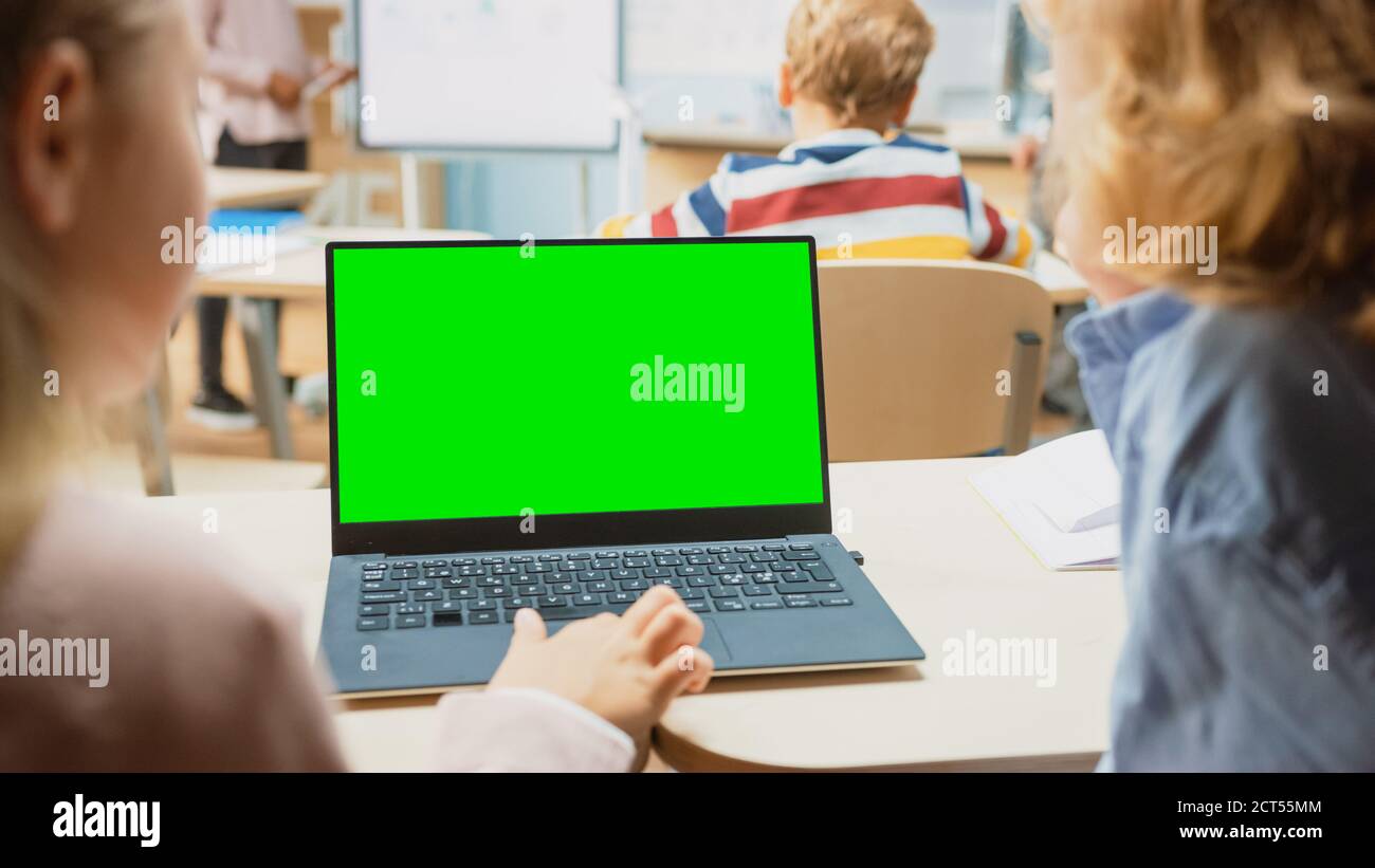 Scuola elementare Classe scientifica: Oltre la spalla Little Boy e Girl utilizzare notebook con schermo verde Mock-up modello su uno schermo. Insegnante di fisica Foto Stock