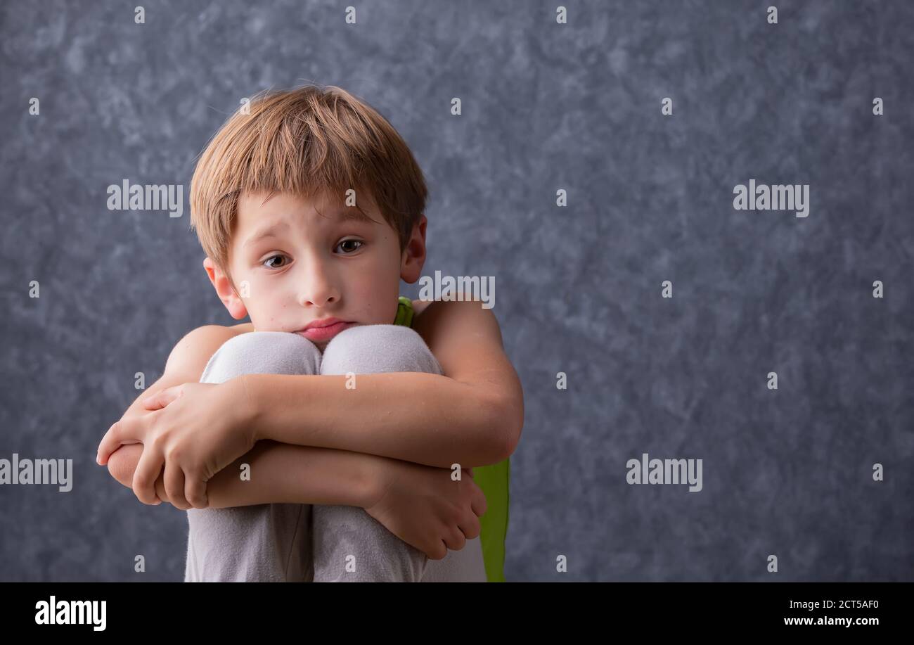 Un bambino triste si siede abbracciando le ginocchia. Un ragazzo di età elementare è triste. Scolaro disunì su uno sfondo grigio. Foto Stock