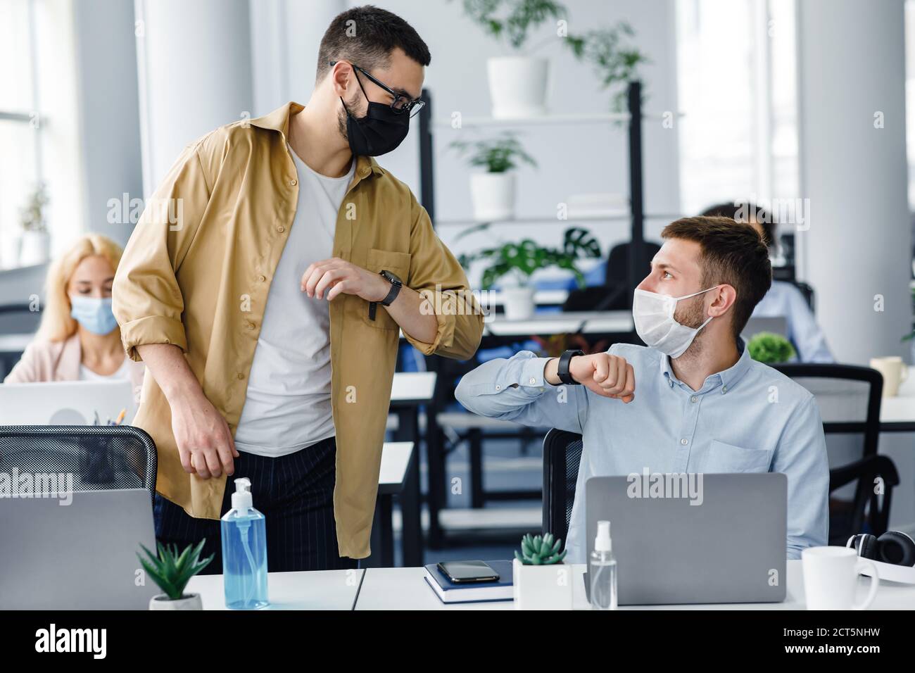 Saluto sicuro al lavoro durante l'epidemia di COVID-19. Giovani uomini in maschere protettive salutano con gomiti sul posto di lavoro con laptop e antisettico in ufficio Foto Stock