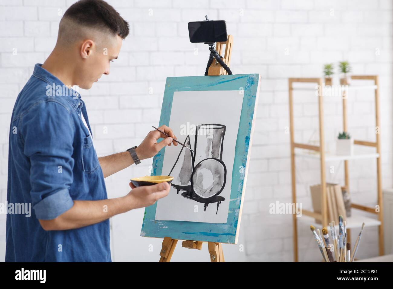 Lezioni di arte moderna e pratica di disegno a casa e isolamento di quarantena. Il giovane uomo in camicia denim disegna la foto sul cavalletto con lo smartphone Foto Stock