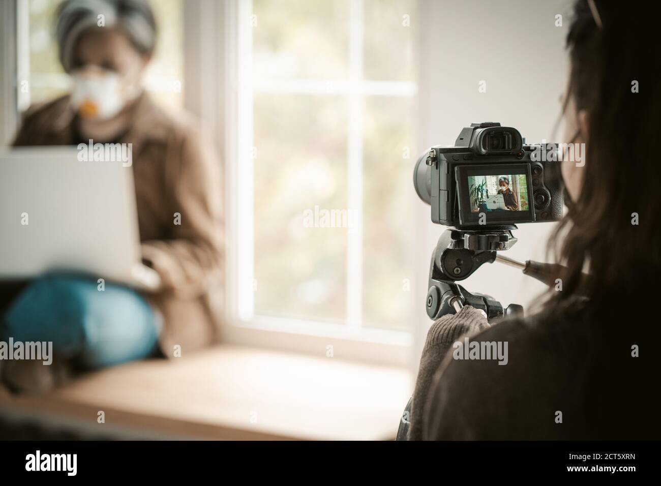 Backstage delle riprese. Messa a fuoco selettiva sulla fotocamera digitale in primo piano. Donna in maschera di faccia con il computer portatile seduto vicino alla finestra su sfondo sfocato Foto Stock