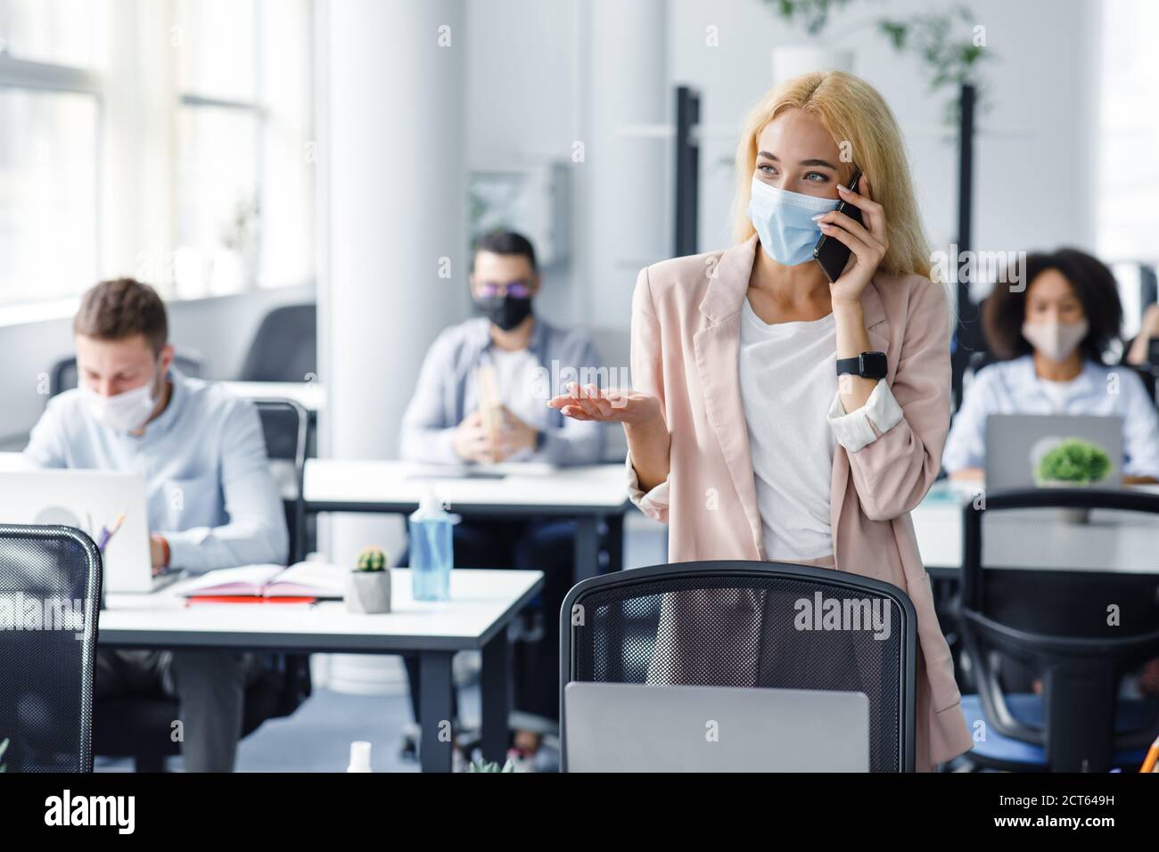 I giovani tornano al lavoro in ufficio dopo il blocco. Donna di affari in maschera protettiva parla al telefono con il cliente Foto Stock
