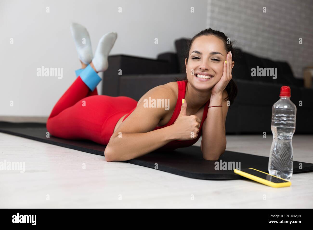 Giovane donna in tuta rossa che fa esercizio o yoga a casa. Ragazza rilassata e ben costruita che giace sul tappetino e mostra il pollice grande in su. Simbolo simile. Riposa dopo Foto Stock