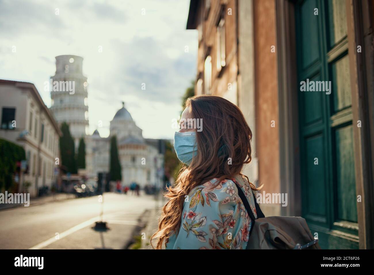 Viaggia durante la pandemia del coronavirus. Donna viaggiatore di mezza età alla moda in abito floreale con maschera medica e zaino godendo della passeggiata vicino a pendente Foto Stock