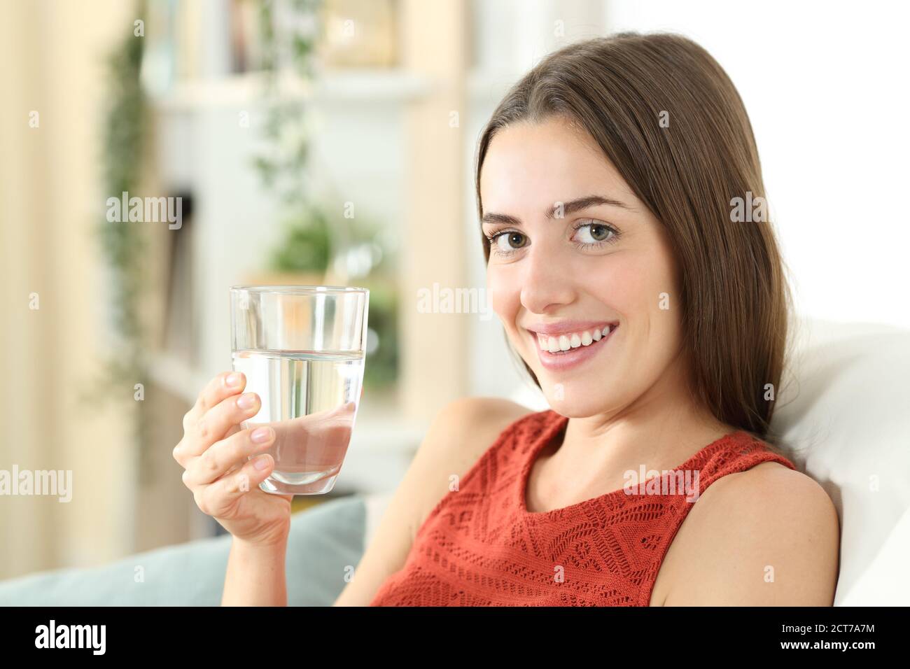La donna felice sta tenendo un vetro di acqua minerale seduto sopra un divano nel soggiorno di casa Foto Stock