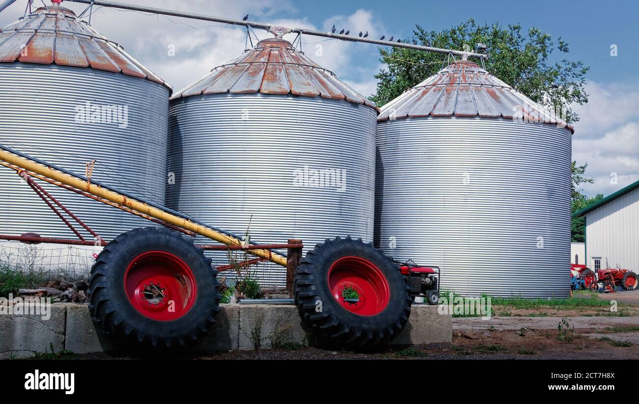 Due pneumatici grandi per trattori con cerchioni rossi luminosi si trovano di fronte a tre silos di grano argentati in un'azienda agricola. Sullo sfondo è presente un trattore rosso brillante Foto Stock