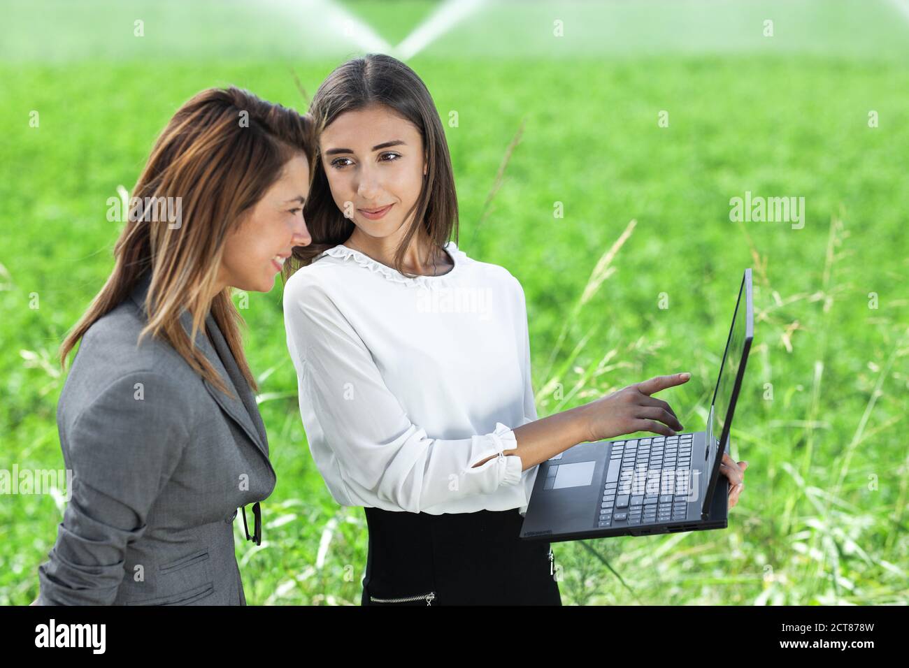 Donne d'affari con un computer portatile in un campo con sistema di irrigazione agricola. Sprinkler ad acqua sullo sfondo. Foto Stock