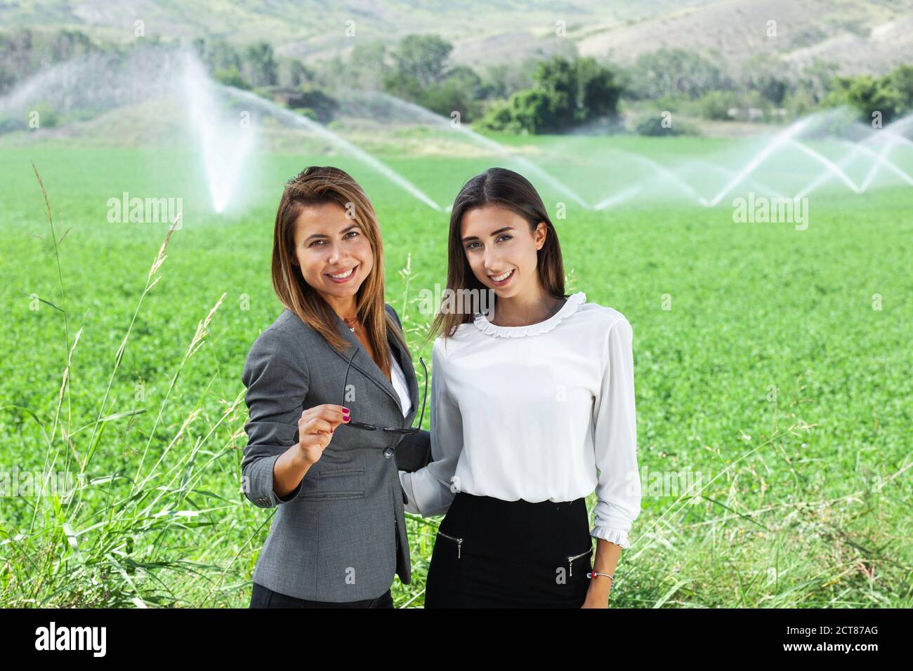 Donne d'affari con un computer portatile in un campo con sistema di irrigazione agricola. Sprinkler ad acqua sullo sfondo. Foto Stock