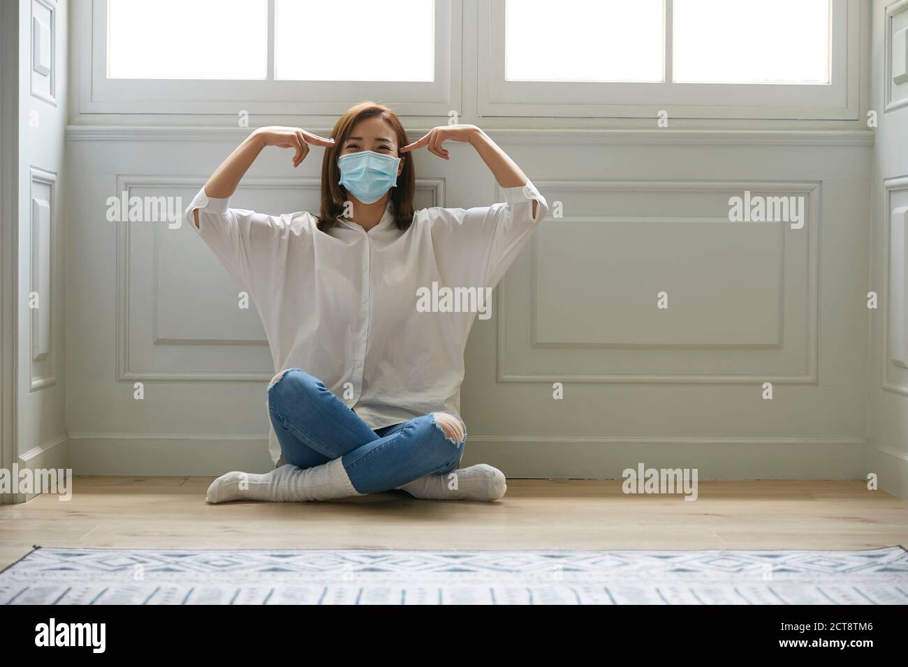 giovane donna asiatica in quarantena a casa con maschera facciale seduto sulle gambe del pavimento attraversato guardare felice Foto Stock