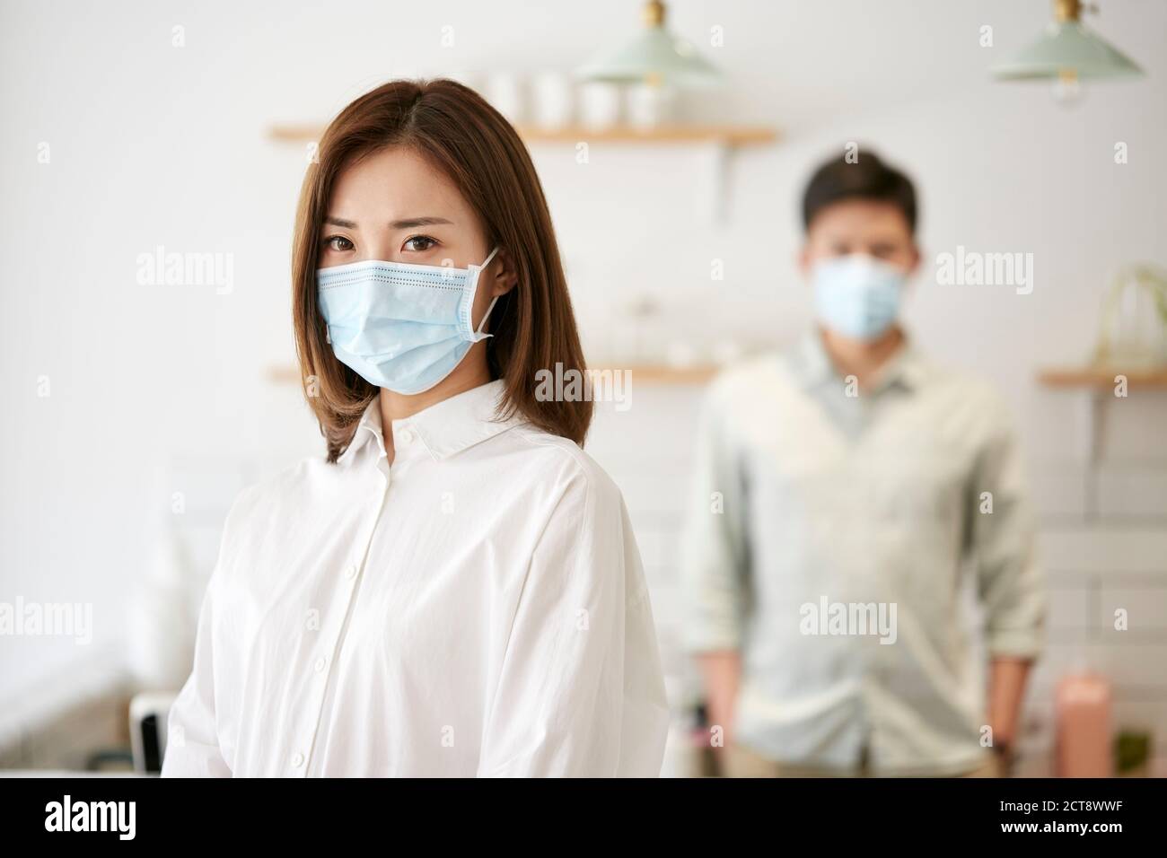 ritratto di una giovane donna asiatica che indossa una maschera guardando fotocamera con moglie sullo sfondo Foto Stock