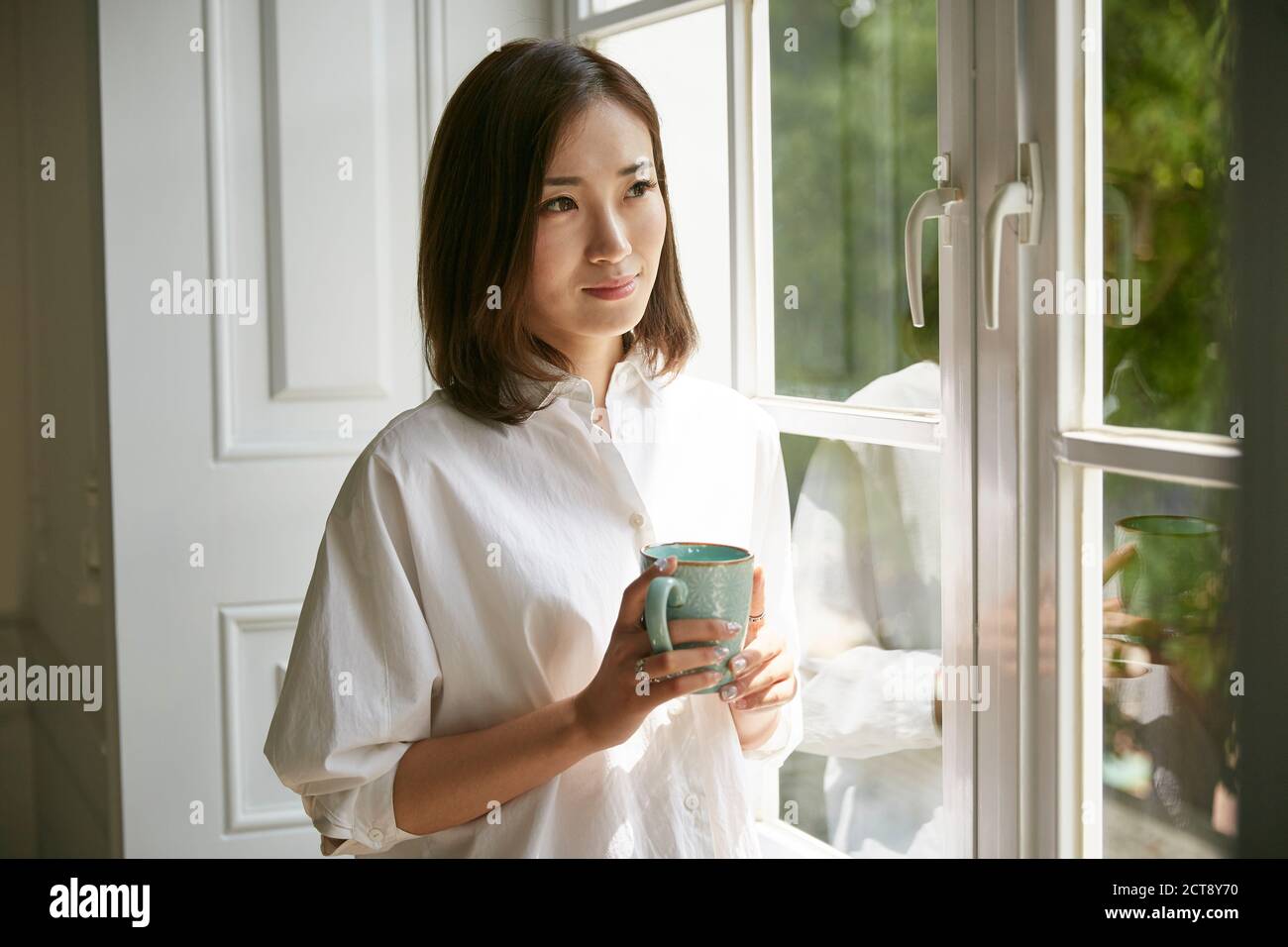 giovane donna asiatica in piedi dalla finestra a casa tenendo un tazza di caffè Foto Stock