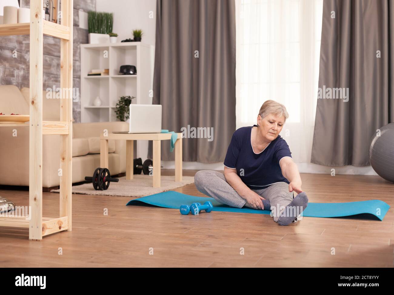 Donna pensionata che si allunga a casa sul tappetino yoga. Anziano pensionato online internet esercizio di formazione a casa attività sportiva con manubri, banda di resistenza, palla svizzera all'età di anziano pensionamento Foto Stock