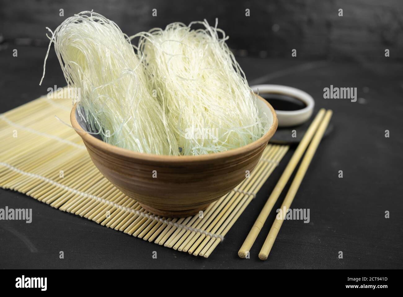 noodle di riso di vetro in una ciotola, fondo asiatico della cucina con stuoia di bambù, chopsticks, salsa di soia su sfondo nero Foto Stock