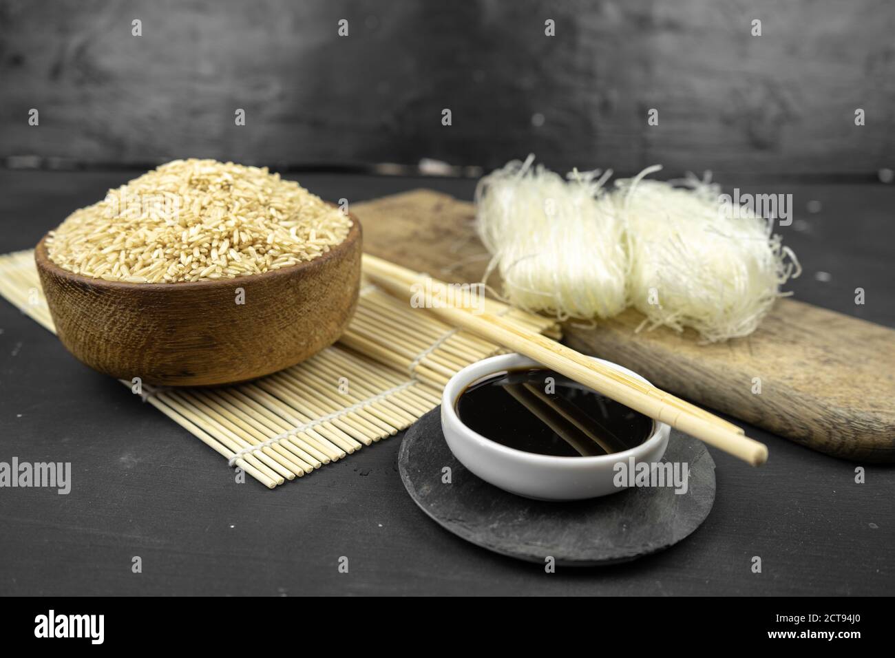 riso marrone in una ciotola di legno, fondo asiatico della cucina con stuoia di bambù, chopsticks, salsa di soia, zenzero sottaceto e spaghetti di riso di vetro su sfondo nero Foto Stock