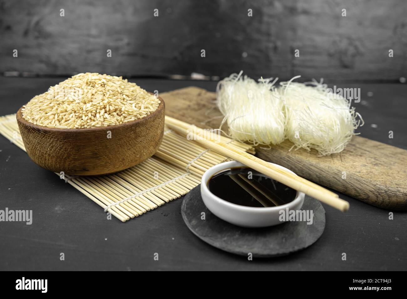 riso marrone in una ciotola di legno, fondo asiatico della cucina con stuoia di bambù, chopsticks, salsa di soia, zenzero sottaceto e spaghetti di riso di vetro su sfondo nero Foto Stock