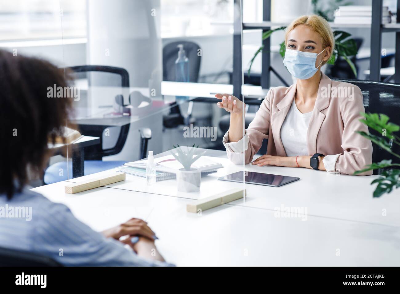 Donna afroamericana parla alla donna d'affari in maschera protettiva attraverso la partizione in vetro all'interno dell'ufficio Foto Stock