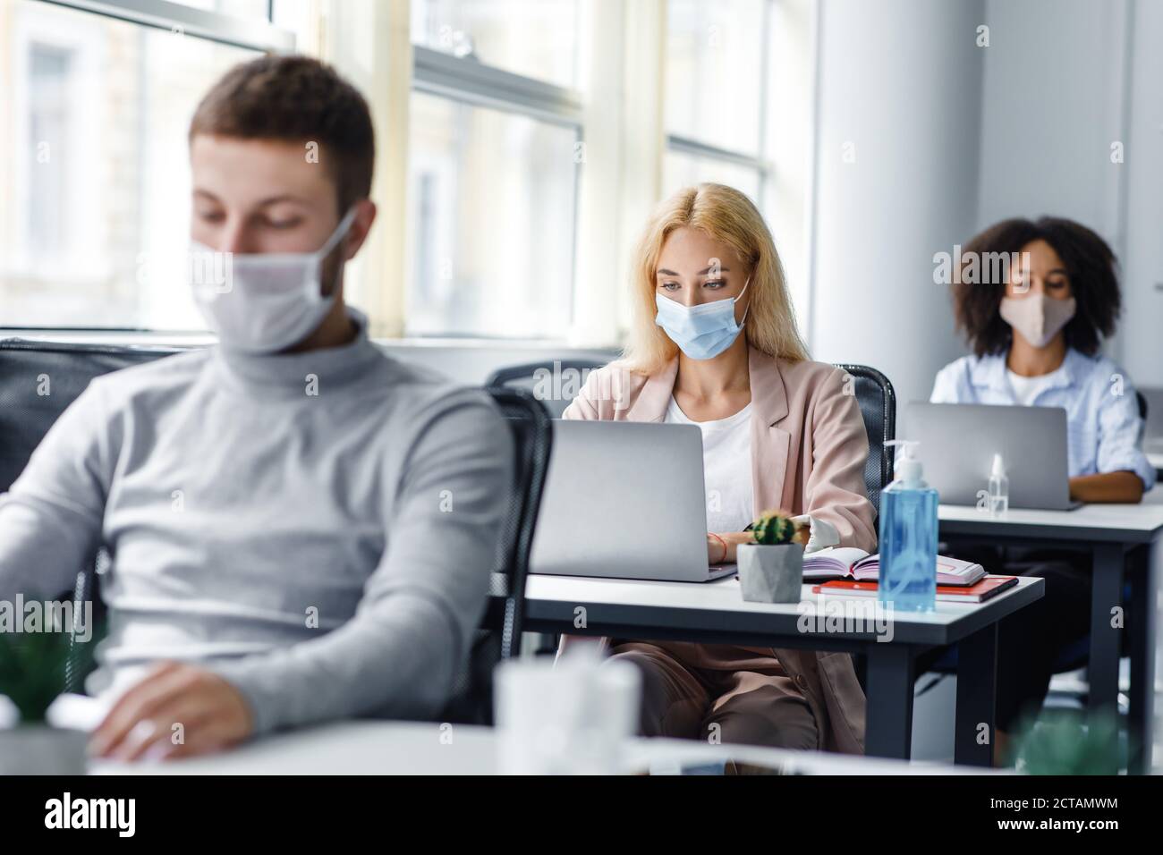 Distanza sociale e nuova normalità sul posto di lavoro. Uomo multirazziale e donne in maschere di protezione guardare i computer portatili a tavoli con antisettici Foto Stock