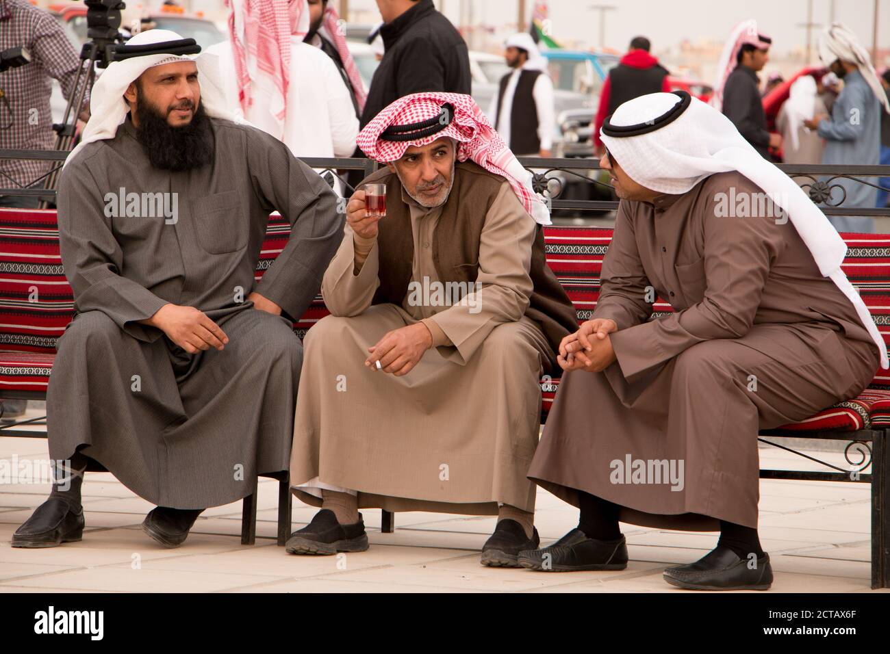 Tre uomini kuwaitiani che parlano e fumano su una panchina. Vestita con piatti tradizionali di colore invernale. Kuwait, Medio Oriente. Foto Stock