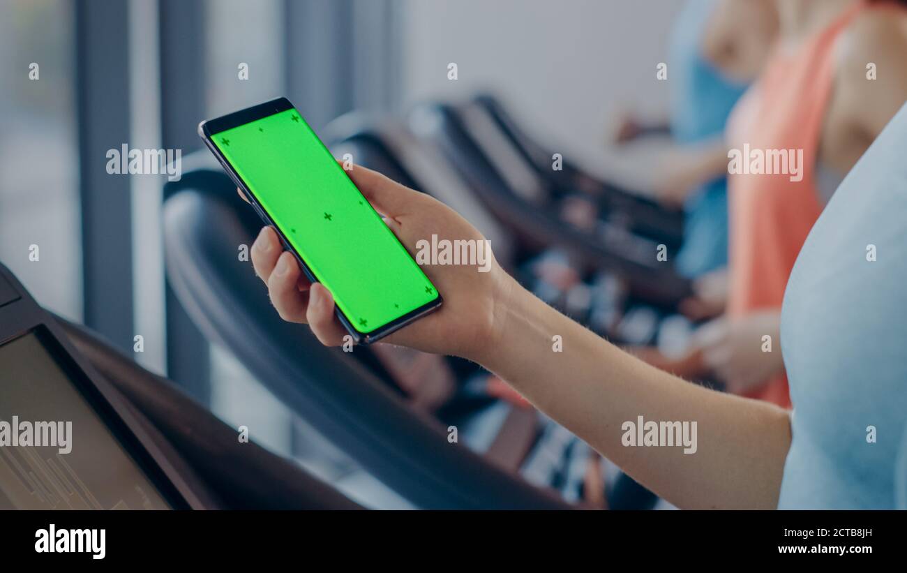 La donna atletica fa gesti mentre si usa lo smartphone Green Screen e si corre sul tapis roulant in una palestra. Fit Female Post sui social media, prende Foto Stock