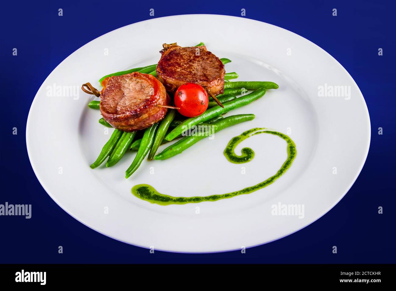 Medaglioni di maiale fritti su spiedini con fagioli e un pomodoro intero. Vista dall'alto su una piastra bianca su sfondo blu. Foto Stock