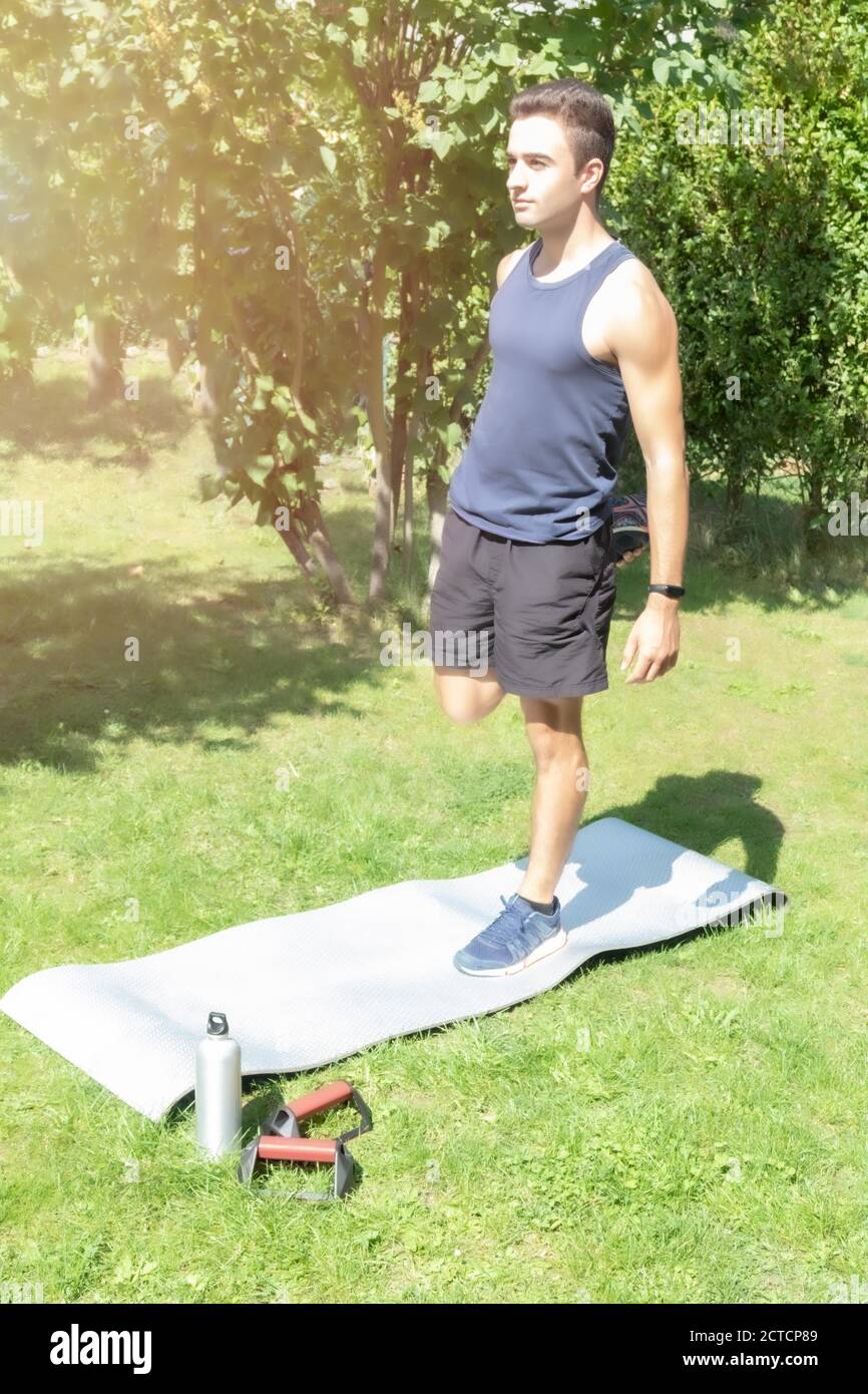 un ragazzo in abbigliamento sportivo in piedi su un tappeto e fare esercizi  di allungamento delle gambe nel suo cortile dove si può vedere una bottiglia  d'acqua e un'attrezzatura per l'addestramento su