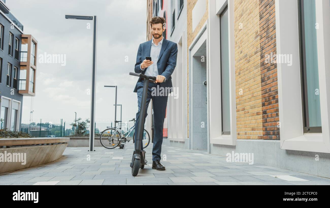 Il giovane uomo d'affari sta verificando il suo smartphone mentre si guida per lavorare su uno scooter elettrico. L'imprenditore moderno usa il trasporto ecologico contemporaneo a. Foto Stock