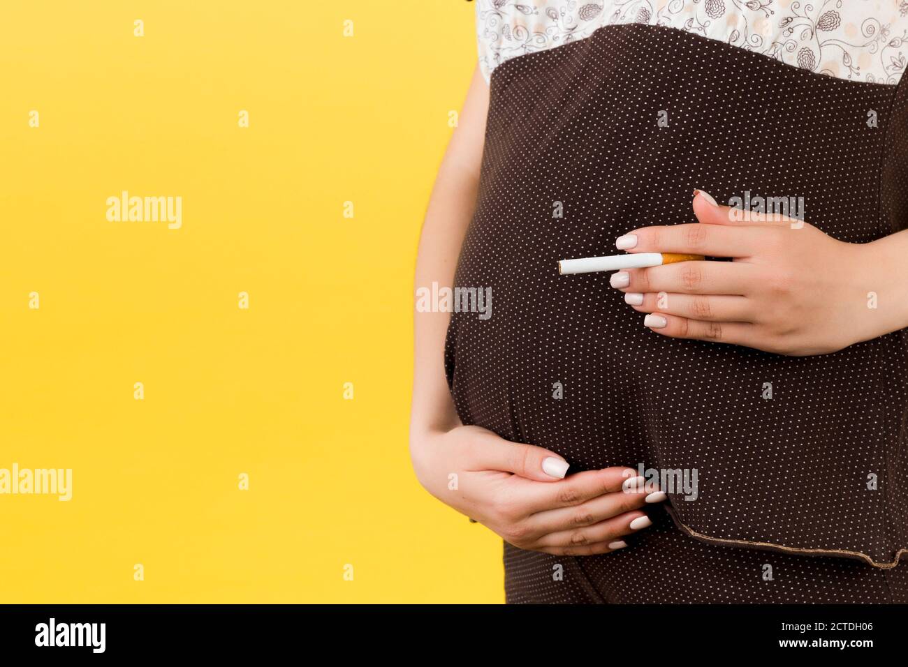 Primo piano di donna incinta in pigiama marrone con una sigaretta contro il ventre su sfondo giallo. Rischio pericoloso per il feto. Foto Stock